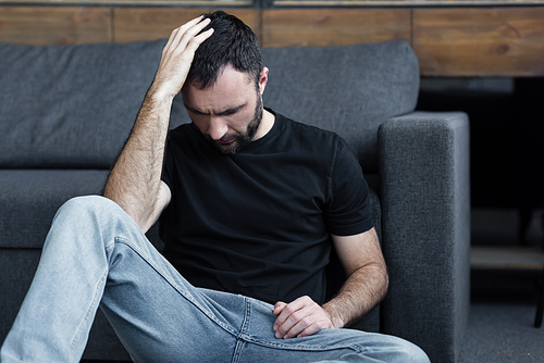 depressed bearded man sitting on floor near grey sofa and holding hand on head