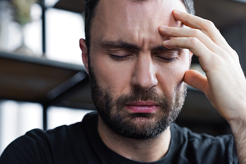 sad unshaven man holding hand near head with closed eyes