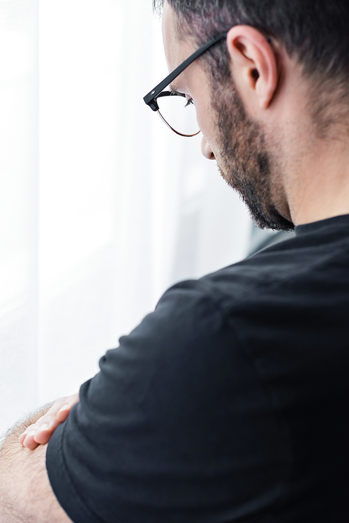 unshaven depressed man in glasses standing by window at home