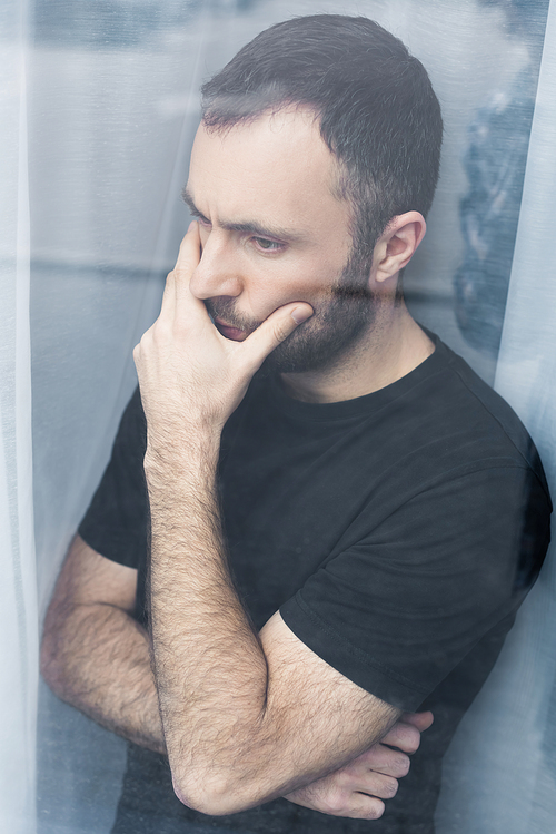 handsome, pensive man in black t-shirt standing by window and holding hand on mouth