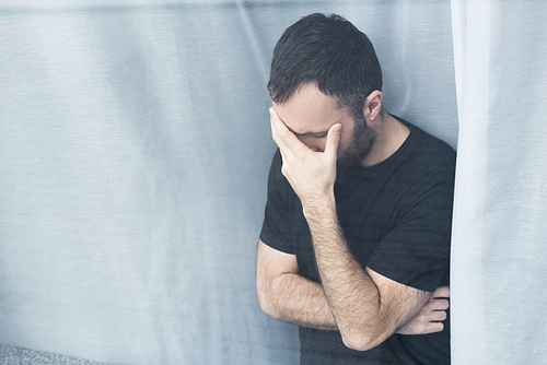 lonely man in black t-shirt standing by window at home and holding hand on face