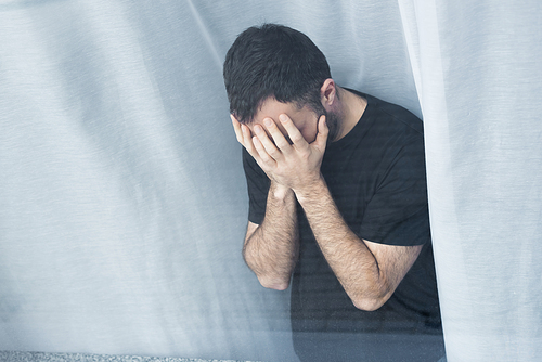 high angle view of lonely man standing by window and holding hands on face