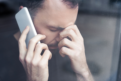 sad handsome man standing by window with smartphone and holding hand near face