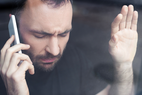 stressed man with closed eyes holding smartphone while standing by window