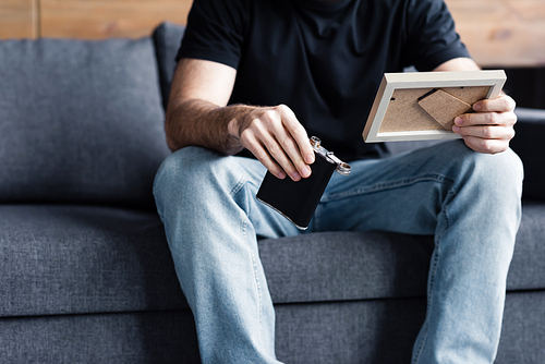 cropped view of man sitting on grey sofa and holding photo frame and flask with alcohol