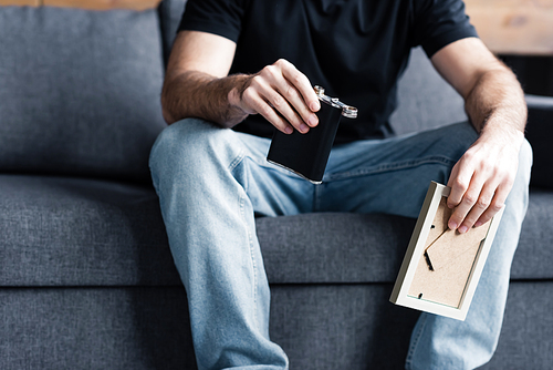 partial view of man sitting on grey sofa and holding photo frame and flask with alcohol