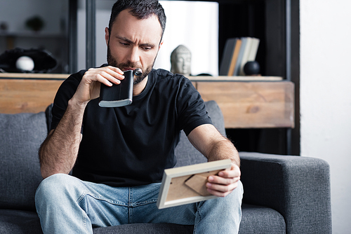 depressed man drinking from flask and holding photo frame while sitting on sofa at home