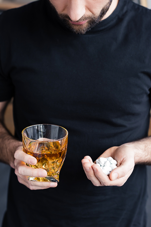 partial view of standing man in black t-shirt holding glass of whiskey and handful of pills