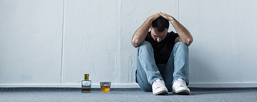 panoramic shot of suffering man sitting on floor by white wall near bottle and glass of whiskey