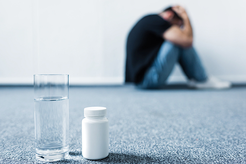 selective focus of depressed man suffering while sitting on floor by wall near container with pills and glass of water
