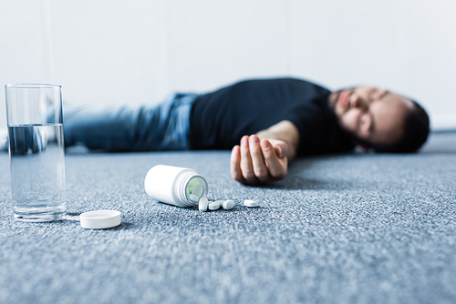 selective focus of unconscious man lying on grey floor near glass of water and container with pills