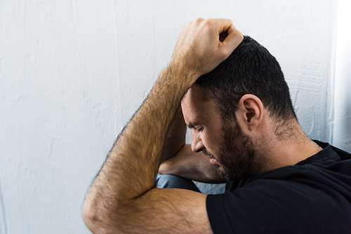 bearded depressed man standing near white wall  and holding hands on head