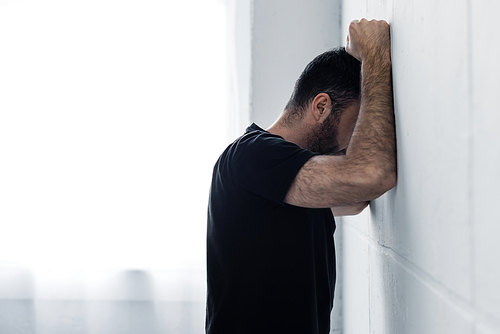 adult depressed man in black t-shirt suffering while standing near white wall