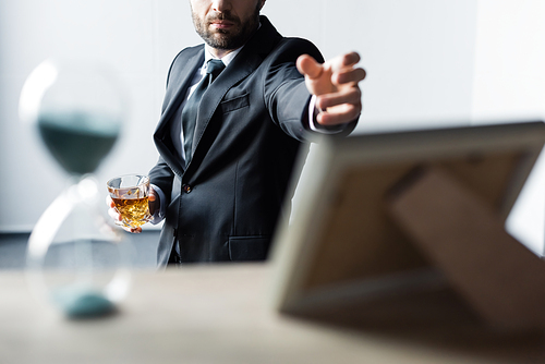 selective focus of man in suit holding glass of whiskey and outstretching hand to table with hourglass and photo frame