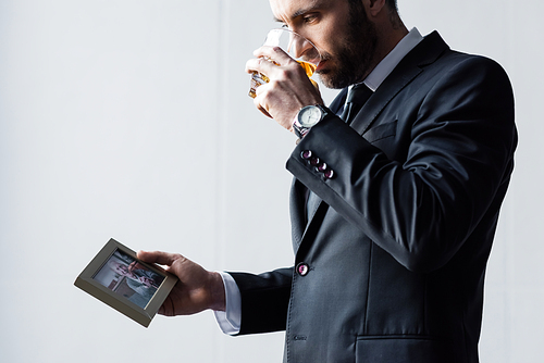 upset man in suit drinking whiskey and looking at photo in frame