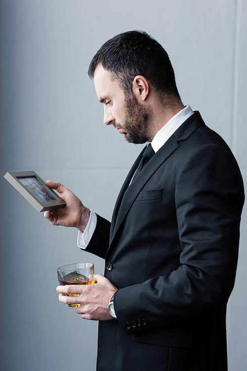 depressed bearded man in suit looking at photo in frame while holding glass of whiskey