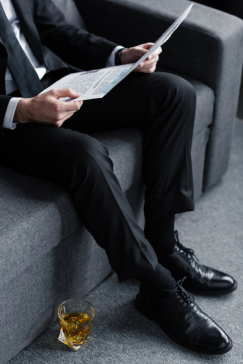 businessman sitting on sofa with newspaper near glass of whiskey on floor