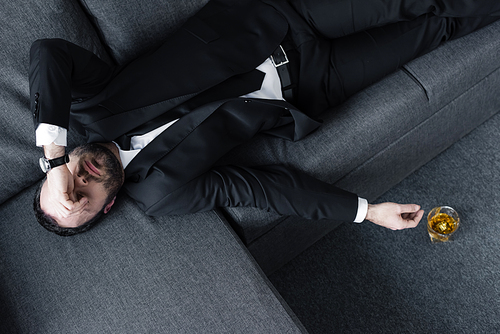 top view of depressed businessman lying on sofa near glass of whiskey on floor
