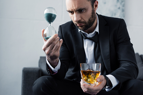 selective focus of upset businessman looking at hourglass while holding glass of whiskey