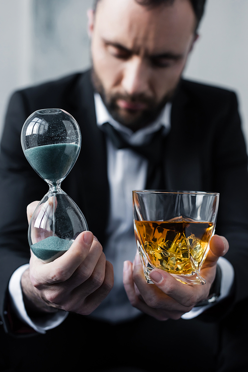 selective focus of upset businessman holding hourglass and glass of whiskey