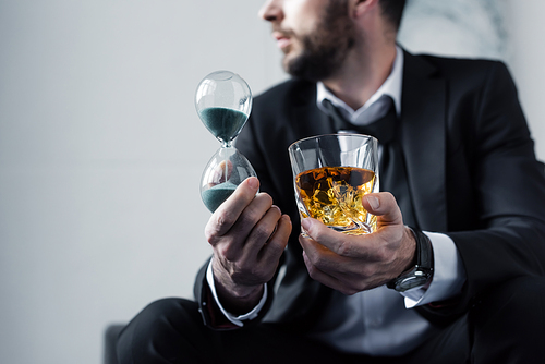 partial view of adult man in suit holding hourglass and glass of whiskey