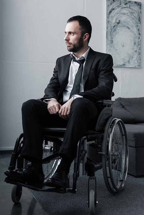 pensive disabled man in black suit sitting in wheelchair and looking away