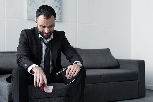 depressed bearded man sitting on sofa while holding gift box and flask