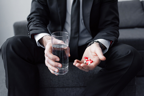 cropped view of man in black suit holding glass of water and handful of pills