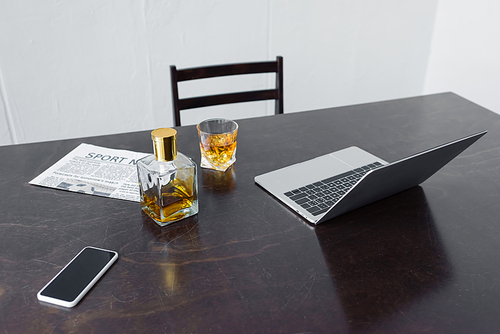 wooden desk with laptop, sport news newspaper, smartphone with blank screen, bottle and glass of whiskey