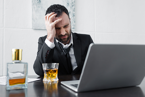 upset businessman sitting at workplace near laptop, bottle and glass of whiskey