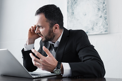 angry businessman quarreling while sitting at workplace and talking on smartphone