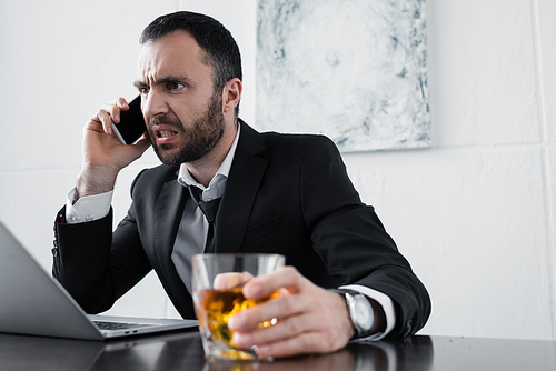 selective focus of angry businessman quarreling while talking on smartphone and holding glass of whiskey
