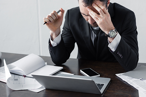 thoughtful businessman sitting at workplace near laptop, documents and smartphone with blank screen