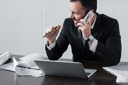 displeased businessman in black suit talking on smartphone while sitting at workplace