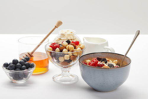 bowls and glasses with fresh berries, nuts, honey and cereal served for breakfast on white table isolated on grey