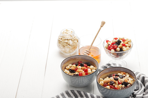 bowls with muesli, dried berries and nuts served for breakfast with honey on white wooden table with copy space
