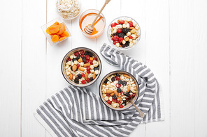 top view of bowls with muesli, dried berries and nuts served for breakfast with dried apricots and honey near striped cloth on white wooden table