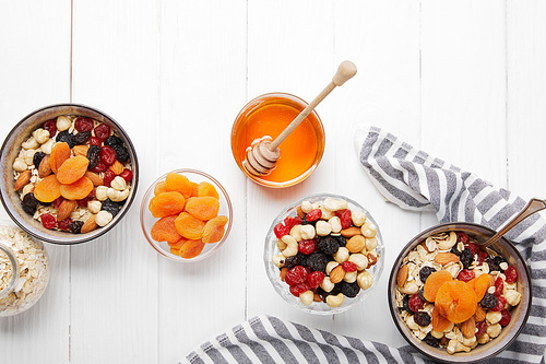 top view of bowls with cereal, dried berries and nuts served for breakfast with dried apricots and honey near striped cloth on white wooden table