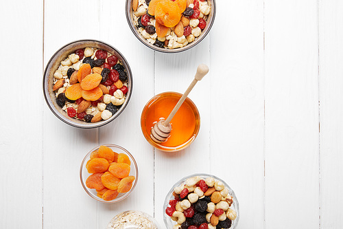 top view of bowls with cereal, dried apricots and berries, honey and nuts on white wooden table