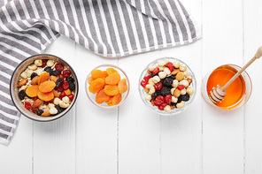 top view of bowls with cereal, dried apricots and berries, honey and nuts on white table with striped cloth