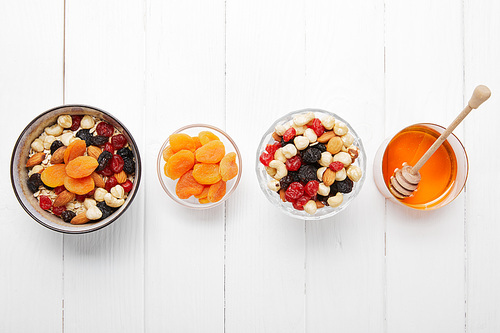 top view of bowls with cereal, dried apricots and berries, honey and nuts in row on white table