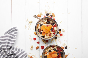 top view of bowls with muesli, dried apricots and berries and nuts served for breakfast with scattered ingredients on white wooden table