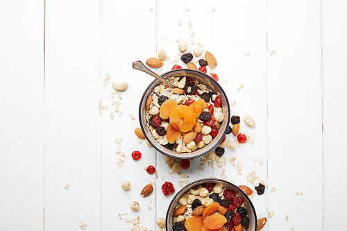 top view of bowls with muesli, dried apricots and berries and nuts served for breakfast with scattered ingredients on white table