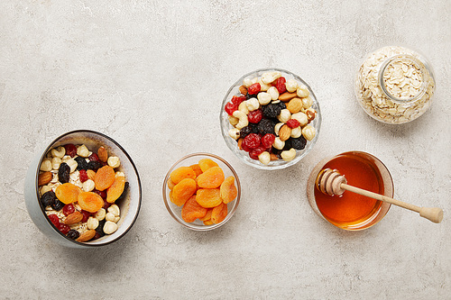 top view of bowls with muesli, dried apricots and berries, nuts and honey on textured grey surface