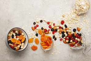 top view of bowls with muesli, dried apricots and berries, nuts on textured grey surface with messy scattered ingredients