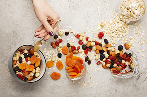 cropped view of woman holding spoon near bowls with muesli, dried apricots and berries, nuts on textured grey surface with messy scattered ingredients