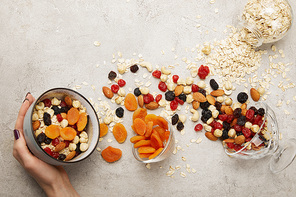 cropped view of woman holding bowl with muesli, dried apricots and berries, nuts on textured grey surface with messy scattered ingredients