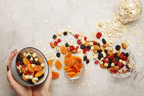 partial view of woman holding bowl with muesli, dried apricots and berries, nuts on textured grey surface with messy scattered ingredients