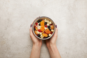 cropped view of woman holding bowl with muesli, dried apricots and berries, nuts on textured grey surface