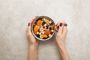 cropped view of woman holding spoon and bowl with muesli, dried apricots and berries, nuts on textured grey surface